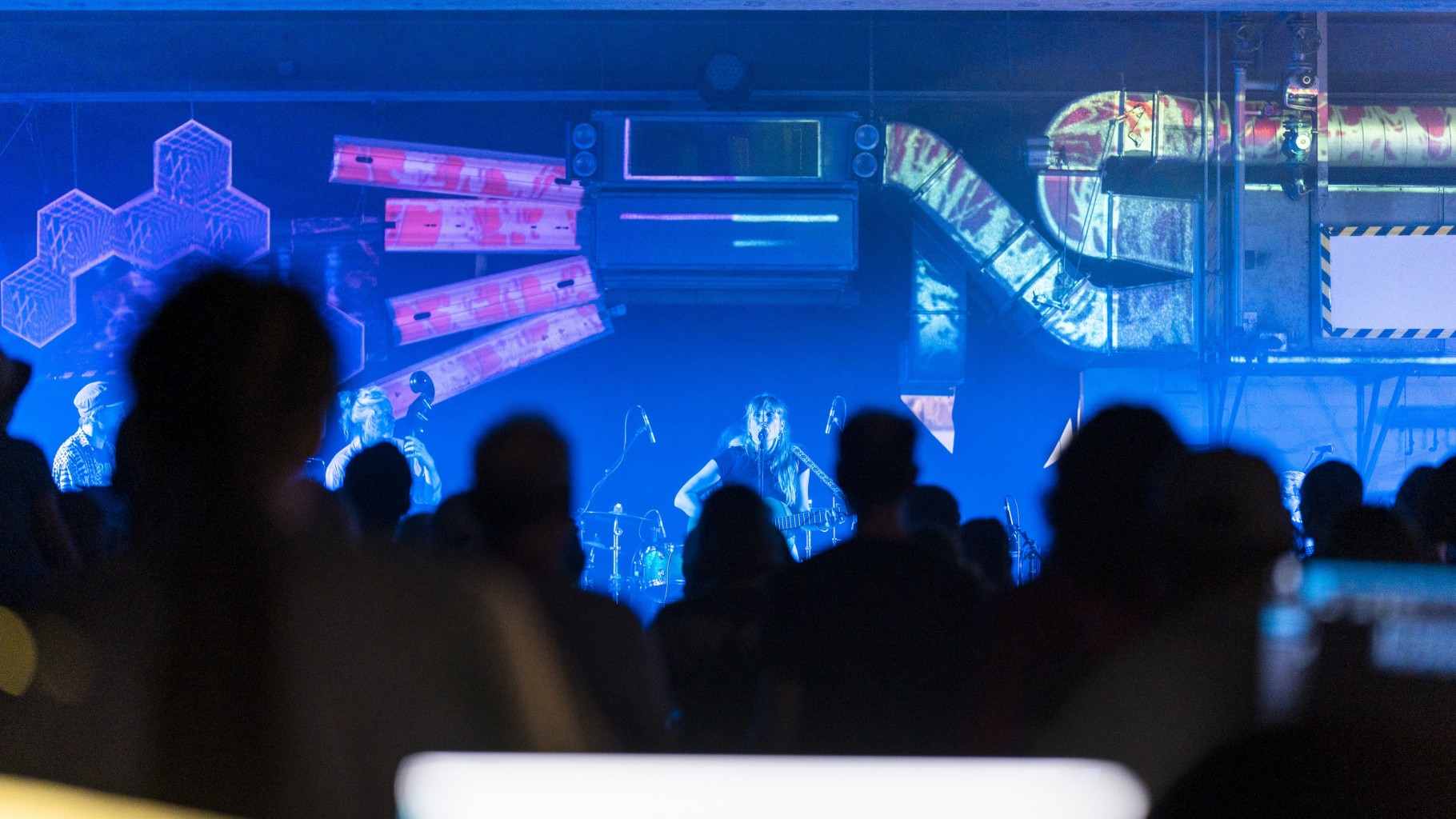 Bild eines Konzerts bei kontakt – dem Kulturfestival 2022; im Vordergrund sind die Silhouetten der Besucher zu sehen. Im Hintergrund spielt gerade eine Band, die Szene ist in blaues Licht getaucht. Auf die Lüftungsschächte und andere Flächen über der Bühne werden bunte Muster projiziert.